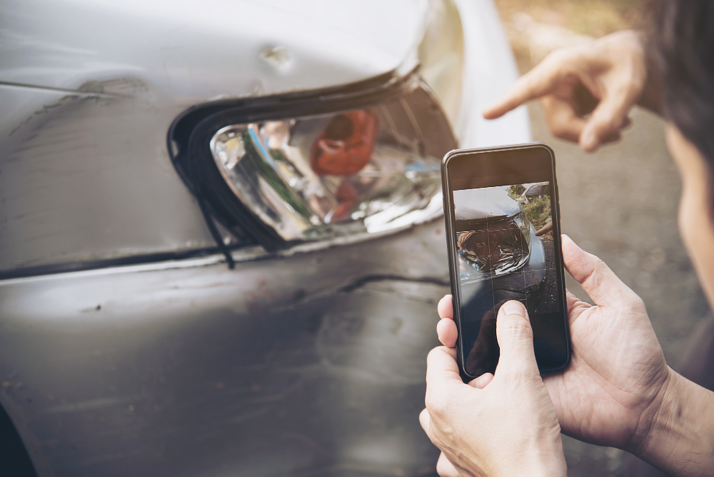 Personne prenant une photo des dommages sur l'épave d'une voiture avec un téléphone portable, tandis qu'une autre personne pointe vers l'impact sur le phare avant